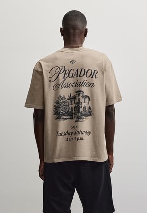 Persoană purtând un tricou bej cu text negru și o ilustrație a unei case pe spate cu inscripția „Pegador Association, deschis marți-sâmbătă 11:00-16:00.”