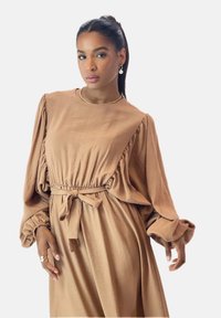 Robe brune fluide avec de longues manches bouffantes, taille froncée et un lien décoratif ; confectionnée en tissu doux, dotée d'un col rond.