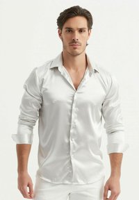 Chemise blanche en satin avec un col pointu, manches longues retroussées et fermeture à boutons sur le devant. Texture lisse et finition brillante.