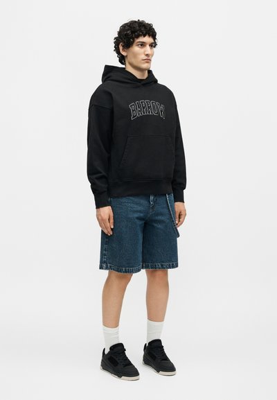 Άντρας που στέκεται φορώντας μαύρο φούτερ με κουκούλα με την επιγραφή "BARROW", μπλε τζιν σορτς, λευκές κάλτσες και μαύρα αθλητικά παπούτσια σε απλό φόντο.