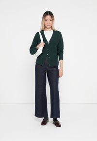Cardigan en tricot vert avec un motif en losanges, boutons sur le devant, associé à un jean large bleu marine et des chaussures sombres, tenant un sac blanc.
