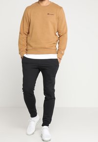 Felpa marrone con logo ricamato, abbinata a pantaloni jogger neri e affusolati. Sneakers bianche completano l'outfit. Design semplice e sportivo.