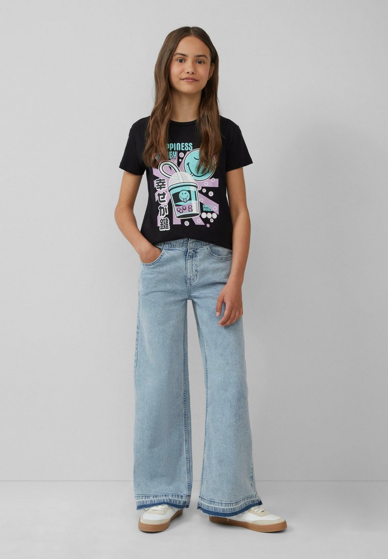 Zwarte graphic t-shirt met kleurrijk ontwerp van een konijn en tekst, gecombineerd met lichtblauwe wijde jeans en witte sneakers.