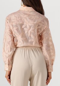 Bluse in Pfirsich mit floralem Stickmuster, transparenter Textur und knöpfbaren Manschetten; getragen über beigen Hosen mit elastischem Bund.