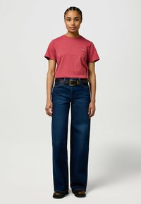 Rode katoenen t-shirt met korte mouwen, met een klein logo; gecombineerd met hoge taille, wijde blauwe spijkerbroek en een bruine riem.