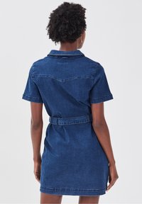 Robe chemise en denim, bleu foncé, taille ceinturée, manches courtes, col, avec des détails de surpiqûres et un empiècement au dos. Texture lisse.