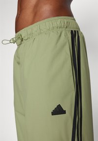 Světlé olivově zelené sportovní kraťasy s elastickým pasem, černými bočními pruhy a vyšitou černou logem na levé nohavici.