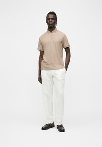 Jeune homme debout, portant un polo beige à manches courtes, un pantalon blanc et des mocassins foncés, une main dans la poche.