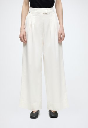 Άτομο που φοράει ψηλόμεσο, φαρδύ λευκό παντελόνι και μαύρα κλειστά, επίπεδα παπούτσια, στέκεται μπροστά σε απλό φόντο.