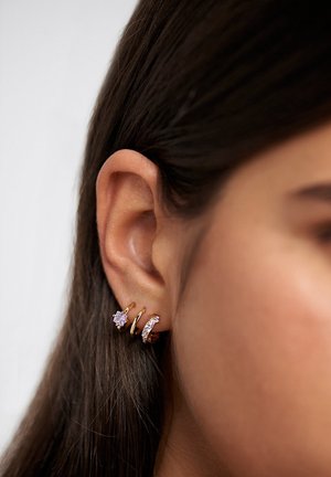 Gros plan sur l'oreille d'une femme ornée de trois petites boucles d'oreilles en or, dont une avec une pierre précieuse en forme d'étoile et une autre incrustée de cristaux.