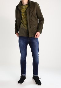 Olivgrön jacka över en gul grafisk t-shirt, ihopparad med mörkblå jeans och svarta sneakers. Slät tygkvalitet med ledig passform.