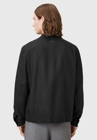 Svart långärmad skjorta med en stilren design och textilstruktur, med rak skärning och en etikett vid baksidan av halsringningen.