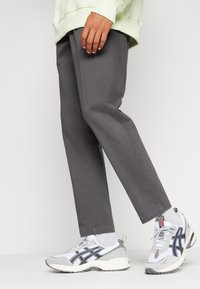 Grijze taps toelopende broek van een lichtgewicht stof, gedragen met witte sportschoenen met grijze en zwarte accenten en gecombineerd met witte sokken.