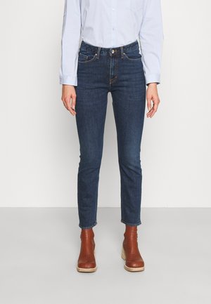 Person iført mørkeblå slim-fit jeans, lyseblå indstøbt skjorte og brune ankelstøvler, der står mod en ensfarvet hvid baggrund.