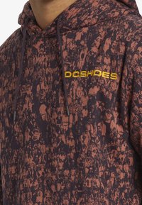 Sudadera con capucha en púrpura oscuro con un patrón de multitudes en marrón rojizo. Cuenta con el logo amarillo de "DC Shoes", cordones y tela texturizada.