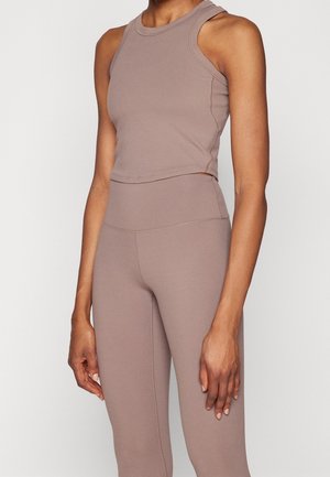 Femme portant un haut court marron sans manches et un legging taille haute assorti sur un fond uni.