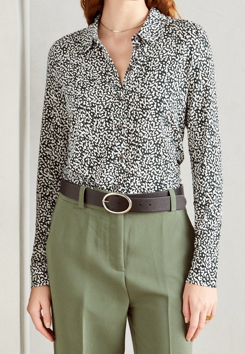 Femme portant une chemise boutonnée noire et blanche à pois, rentrée dans un pantalon taille haute vert olive avec une ceinture noire à boucle ovale.