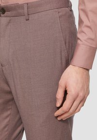Hellbraune maßgeschneiderte Hose mit strukturierter Oberfläche, ausgestattet mit Seitentaschen und einem Knopfverschluss, präsentiert neben einem rosa Langarmhemd.