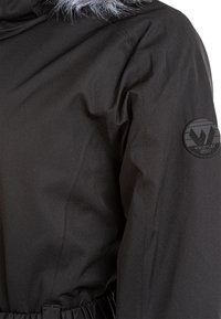 Svart vinterjacka med huva som har grått fuskpälsfoder, elastisk midja och en svart cirkulär logotyp på vänster ärm.