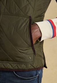 Gepolsterte olivgrüne Weste mit braunen Corduroy-Akzenten, ausgestattet mit einer seitlichen Tasche und einem dezent gestickten Logo nahe dem Saum.