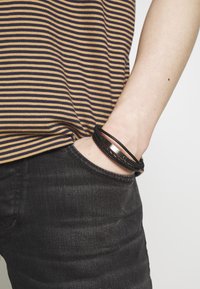 Bracelet en cuir noir et métal avec des textures tressées et lisses, porté au poignet à côté d'une chemise rayée jaune et marine.