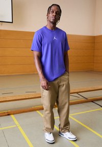 Camiseta de algodón morada con un logo Jumpman blanco, pantalones holgados beige y zapatillas blancas con detalles grises en un gimnasio.