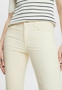 Femme portant un pantalon en velours côtelé crème et un haut blanc à manches courtes avec des rayures horizontales noires, les mains détendues le long du corps.