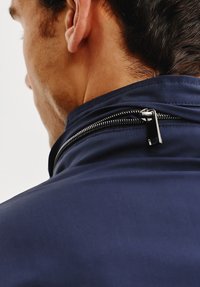 Granatowa kurtka z wysokim kołnierzem i metalowym zamkiem błyskawicznym z tyłu, prezentująca gładką, matową fakturę tkaniny.