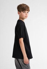 Zwarte katoenen T-shirt met korte mouwen, ronde halslijn en een reguliere pasvorm. Grijze jeans zichtbaar, casual gedragen, tegen een plain witte achtergrond.