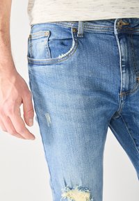 Gros plan d'une personne portant un jean en denim bleu clair déchiré et une chemise beige clair, la main gauche détendue le long du corps.