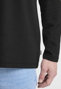 Black long-sleeve shirt made from cotton, featuring a straight cut and a small tag on the left side. Paired with light blue denim.