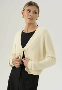 Donna con capelli biondi che indossa orecchini a cerchio dorati, annoda un cardigan color crema sopra una maglietta nera e pantaloni neri, guardando di lato.