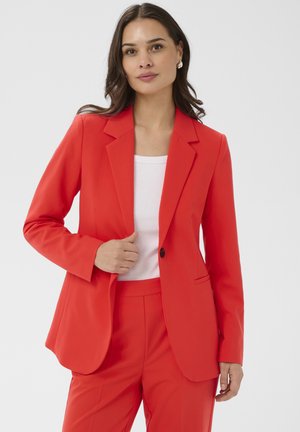 Mujer con cabello castaño que lleva una chaqueta y pantalones rojos, sujetando la solapa de la chaqueta, con una camiseta blanca debajo, sobre un fondo claro y liso.