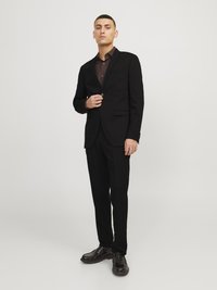 Black suit with a single-button blazer, matching trousers, and a dark brown button-up shirt. Smooth fabric, tailored fit, and polished shoes.