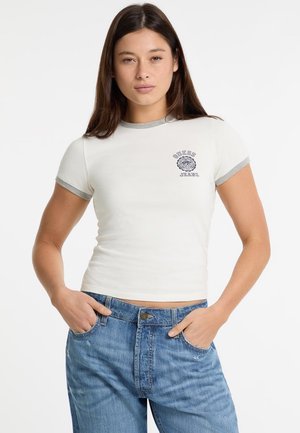 Weißes T-Shirt mit kurzen Ärmeln und grauem Saum, mit einem runden "Guess Jeans"-Logo auf der Brust, kombiniert mit blauen hochgeschnittenen Jeans.