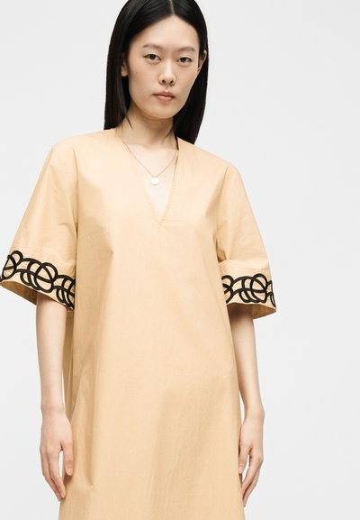 Femme portant une robe beige ample avec une broderie circulaire noire sur les manches, debout devant un fond clair uni.