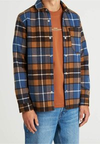 Koszula z flaneli w kratę w kolorach niebieskim, brązowym i czarnym z białymi akcentami. Posiada zatrzaski i kieszeń na piersi; noszona na pomarańczowej koszulce.