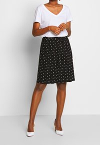 Femme portant un haut blanc à manches courtes et col en V, une jupe noire longueur genou à pois blancs, et des chaussures à talons hauts blanches, debout.