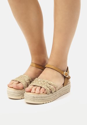 Sandalias de espadrilles tejidas en beige con una plataforma texturizada y una correa marrón en el tobillo, con puntas descubiertas y un diseño trenzado.