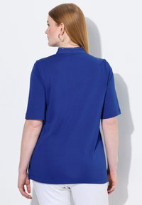 Modrá mikina s krátkým rukávem vyrobená z hladkého materiálu, s vysokým límcem ozdobeným kovovými prvky vzadu a volným střihem.