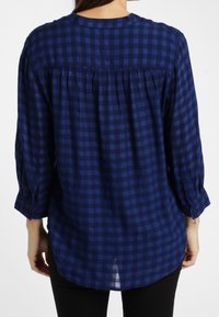 Kvinna iförd en lös, mörkblå och svart rutad långärmad skjorta, sedd bakifrån, ihop med svarta byxor.