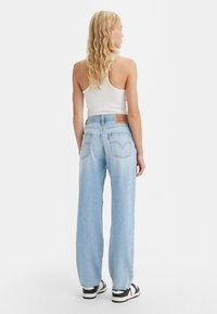 Vaalea nainen, jolla on laineikkaat hiukset, pukeutuneena valkoiseen toppiin, vaaleansinisiin korkeavyötäröisiin farkkuihin ja musta-valkoisiin lenkkareihin, seisoo selin.