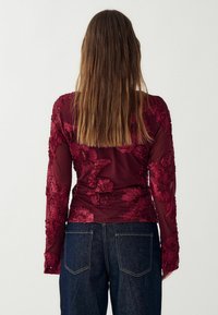 Burgundia pikaevarüstusega top, millel on lilleneedus läbipaistval kangal, koos tumedate denimpükste jahtiga. Seljal on tekstuurilised detailid.