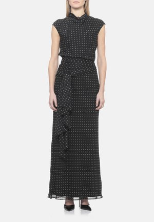 Robe maxi noire sans manches à pois blancs, col montant, taille froncée, détail volant sur le côté, associée à des talons noirs pointus.