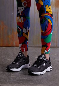 Ténis de desporto pretos com detalhes brancos, tecido texturizado e atacadores. Leggings coloridas com padrões variados de plantas e letras.