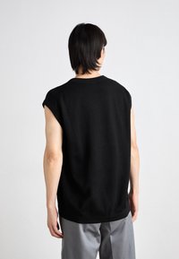 Czarny top bez rękawów z okrągłym dekoltem, posiadający teksturowane boczne panele, luźny krój oraz gładki materiał. Noszony z jasnoszarymi spodniami.