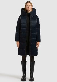 Giacca puffer blu navy con colletto alto, finitura lucida, chiusura con zip e maniche lunghe, che arriva fino a metà coscia. Indossata sopra un outfit nero.