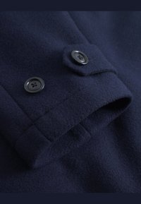 Tela de lana azul marino con una textura suave, que presenta un detalle de puño abotonado. Dos botones negros visibles en la zona de la muñeca.