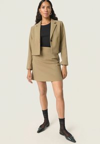 Khaki blézer, cropped fazonnal és egy gombbal, párosítva egy illő szoknyával. Fekete bordázott felső, fekete átlátszó zoknik és hegyes orrú lapos cipők.
