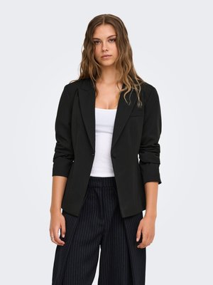 Jonge vrouw met golvend haar, die een zwarte blazer, een wit hemdje en zwarte, wijde broek met fijne krijtstrepen draagt, staand tegen een effen achtergrond.